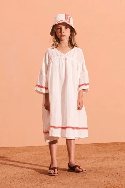 Young girl in white dress and bucket hat poses against peach background. Casual children's fashion shot with warm tones and simple styling.