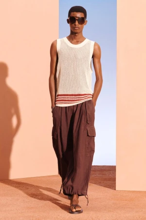 A male model poses in a studio wearing a white tank top, brown cargo pants, and sunglasses against a split peach and blue background.