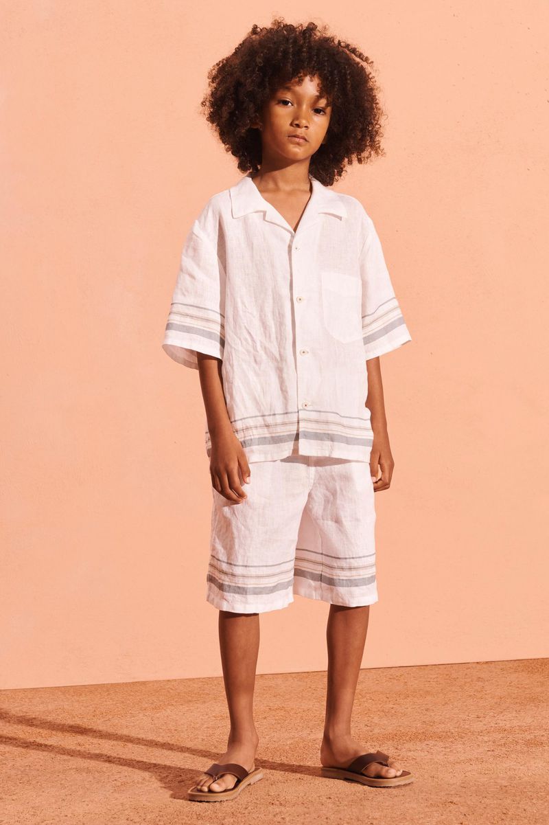 Young boy with curly hair wearing a white linen short set and sandals stands in front of an orange background in a fashion portrait.