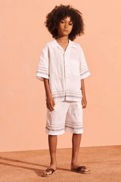 Young boy with curly hair wearing a white linen short set and sandals stands in front of an orange background in a fashion portrait.