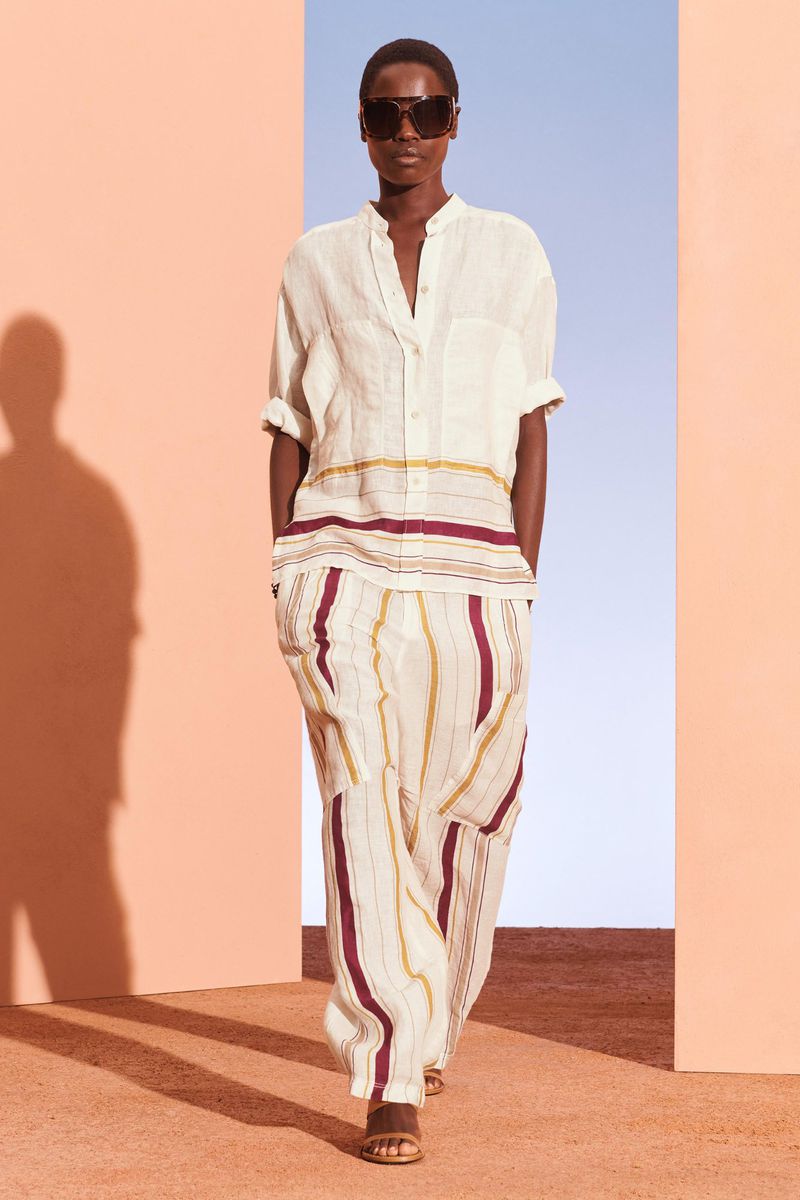 A tanned-skinned woman in a cream-colored striped linen shirt and pants poses indoors with a peach and blue background.