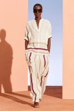 A tanned-skinned woman in a cream-colored striped linen shirt and pants poses indoors with a peach and blue background.