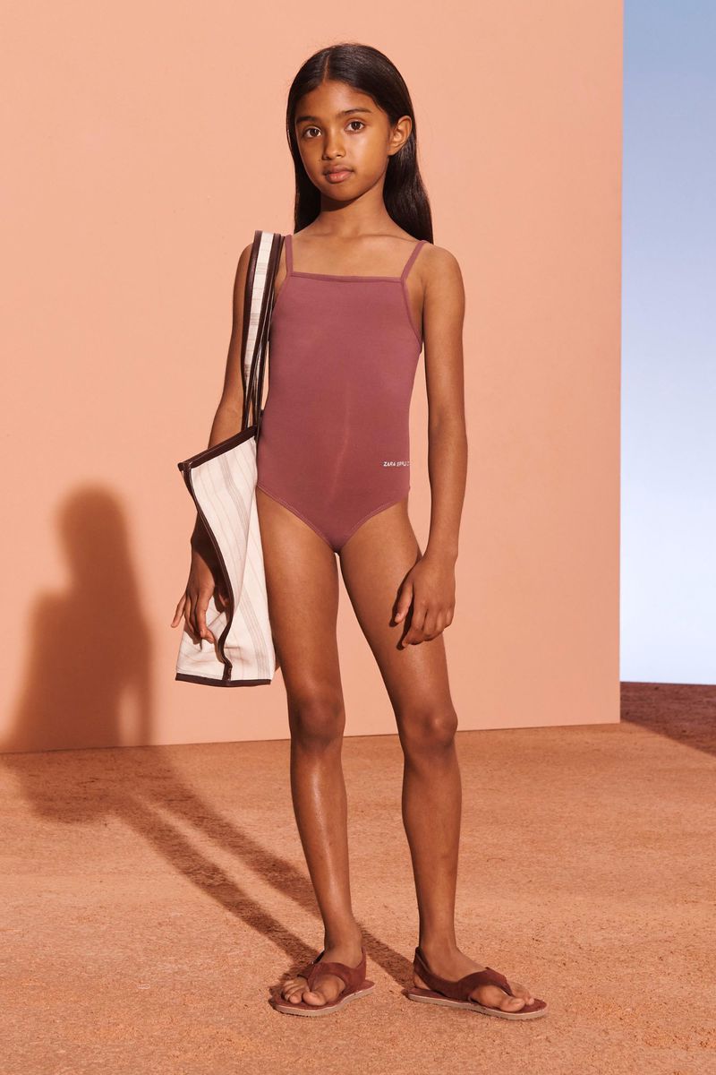 Young child poses in a mauve swimsuit with a white tote bag and brown sandals against a peach and light blue background.