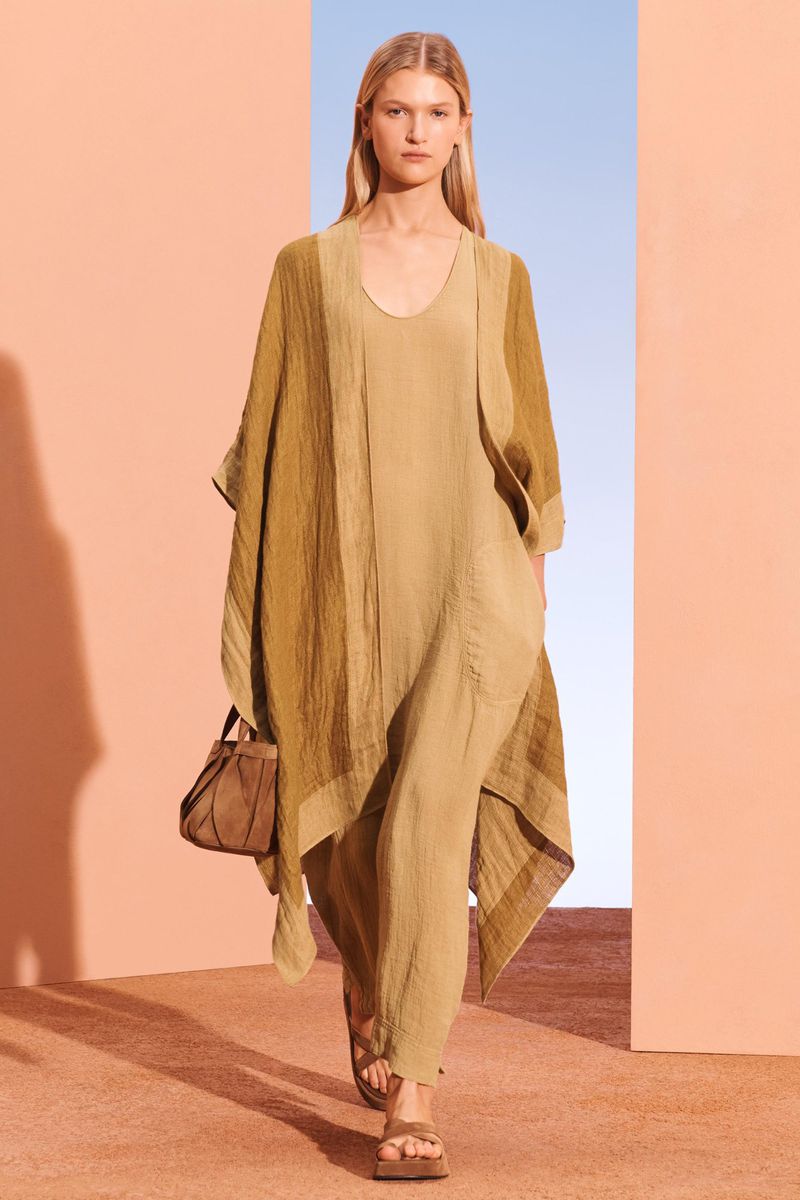 Fashion shot of a blond adult female walking in a tan dress and sandals, carrying a matching purse, against peach and blue background.