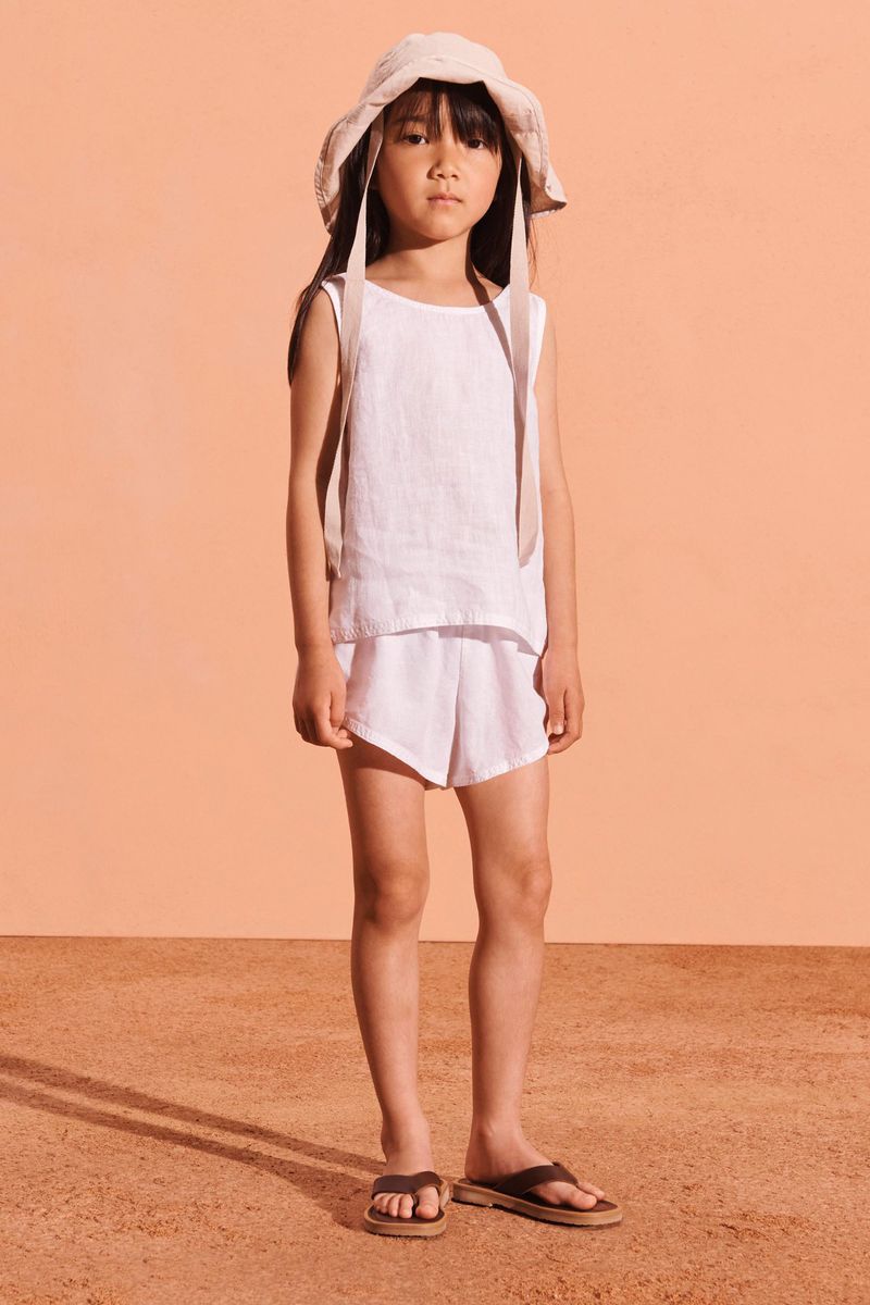 A young girl stands in a studio wearing a white linen outfit with a sun hat, gazing directly at the camera with a neutral expression.
