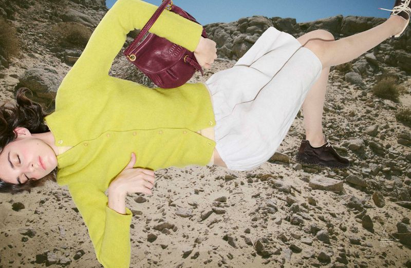 Model wearing a yellow cardigan and white shorts lying on a rocky terrain holding a burgundy handbag
