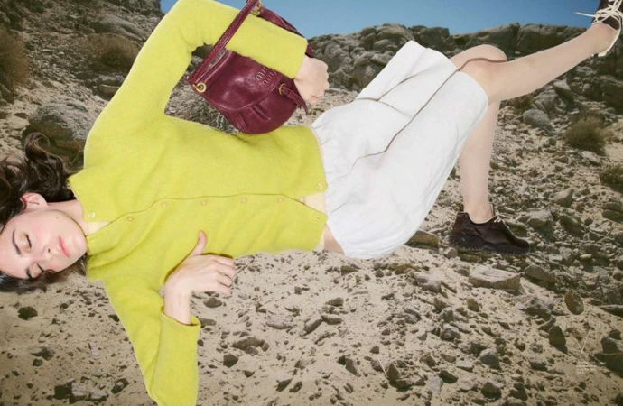 Model wearing a yellow cardigan and white shorts lying on a rocky terrain holding a burgundy handbag