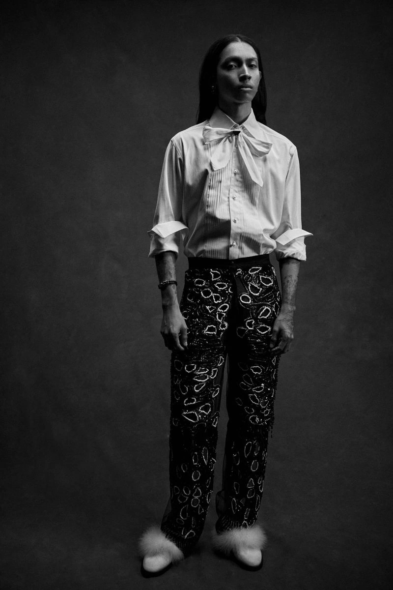Model with long dark hair wearing a white collared shirt with a bow and patterned trousers against a dark backdrop
