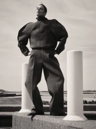 Model Beauïse Genç posing outdoors in structured dark clothing between two white pillars against an overcast sky