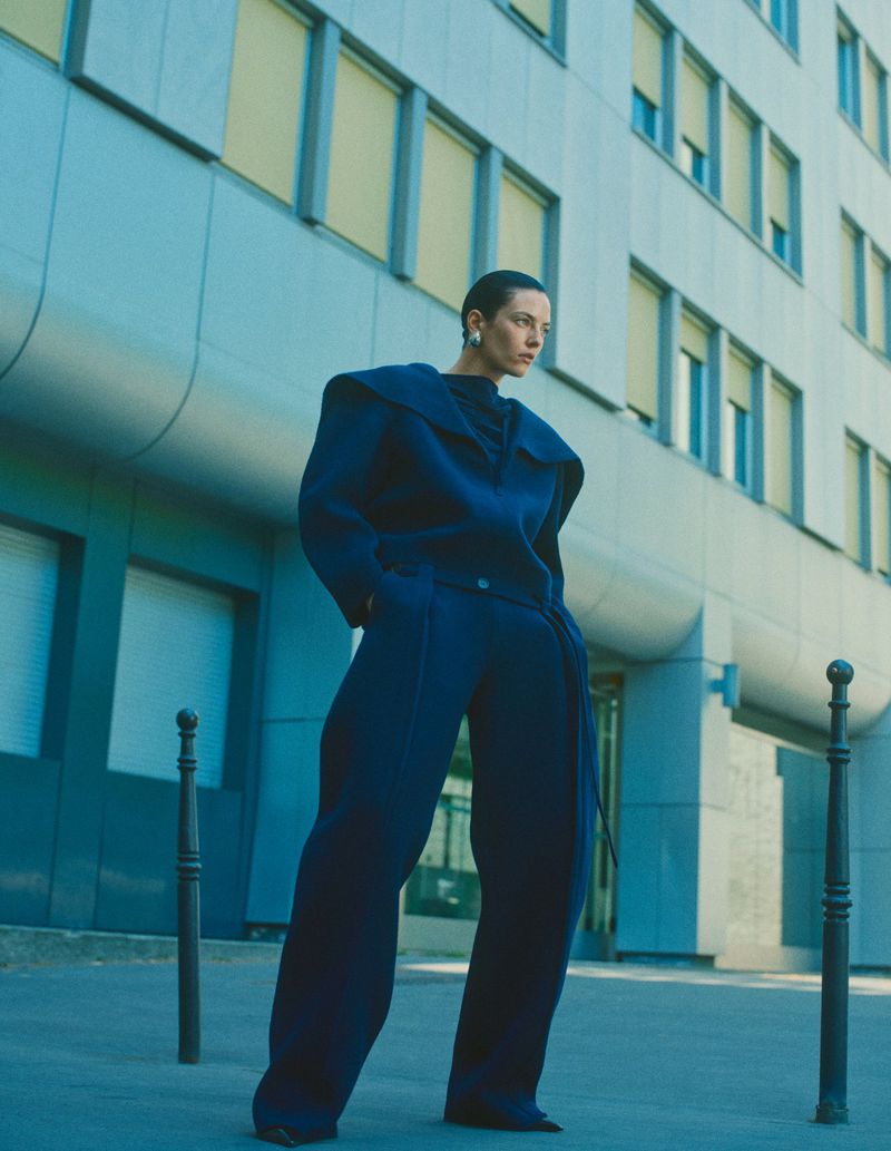 Model Beauïse Genç in a structured blue outfit posing outdoors in front of a modern building exterior.