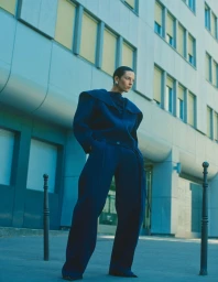 Model Beauïse Genç in a structured blue outfit posing outdoors in front of a modern building exterior.