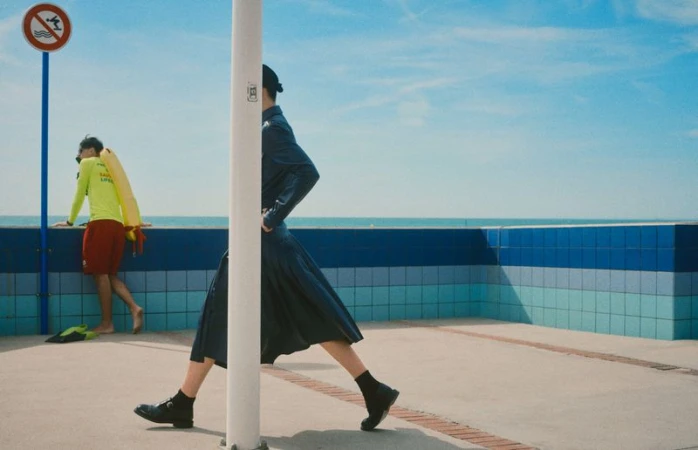 Model in dark skirt and jacket striding past a white pole outdoors near a blue tiled wall and the ocean under a clear sky.