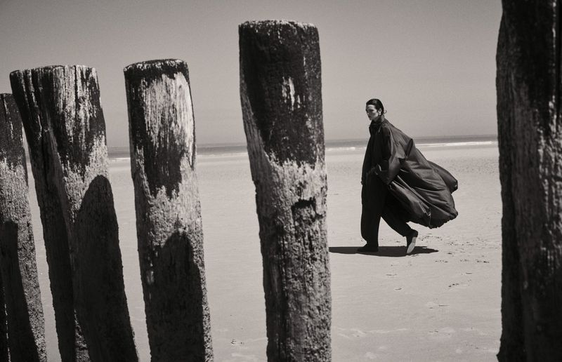 Model walking on a sandy beach past weathered wooden pylons, wearing a flowing dark coat for Elle France.