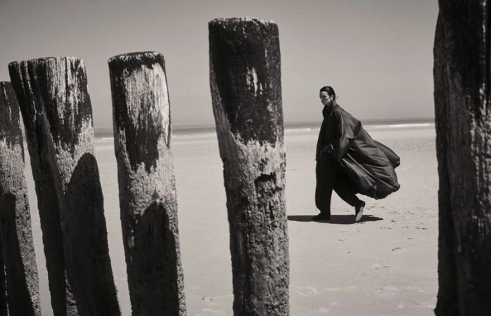 Model walking on a sandy beach past weathered wooden pylons, wearing a flowing dark coat for Elle France.