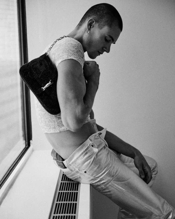 Model Samuel Watson seated by a window holding a dark shoulder bag, wearing light trousers and a cropped patterned top.