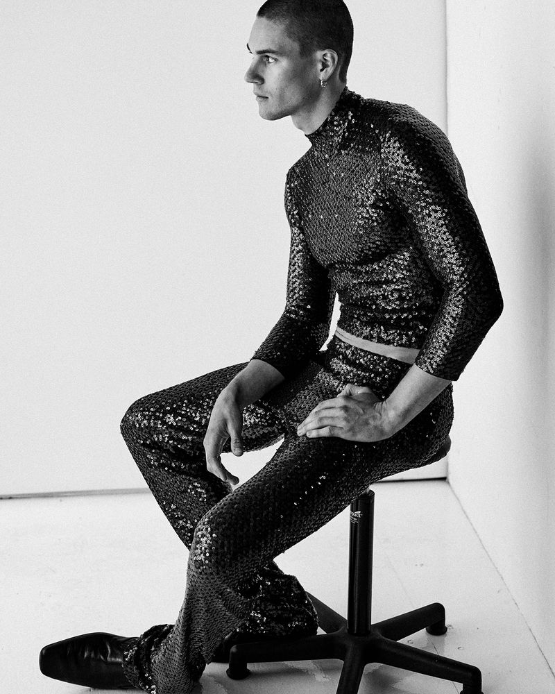 Model Samuel Watson seated on a stool wearing a sequined top and trousers in a monochrome studio portrait.