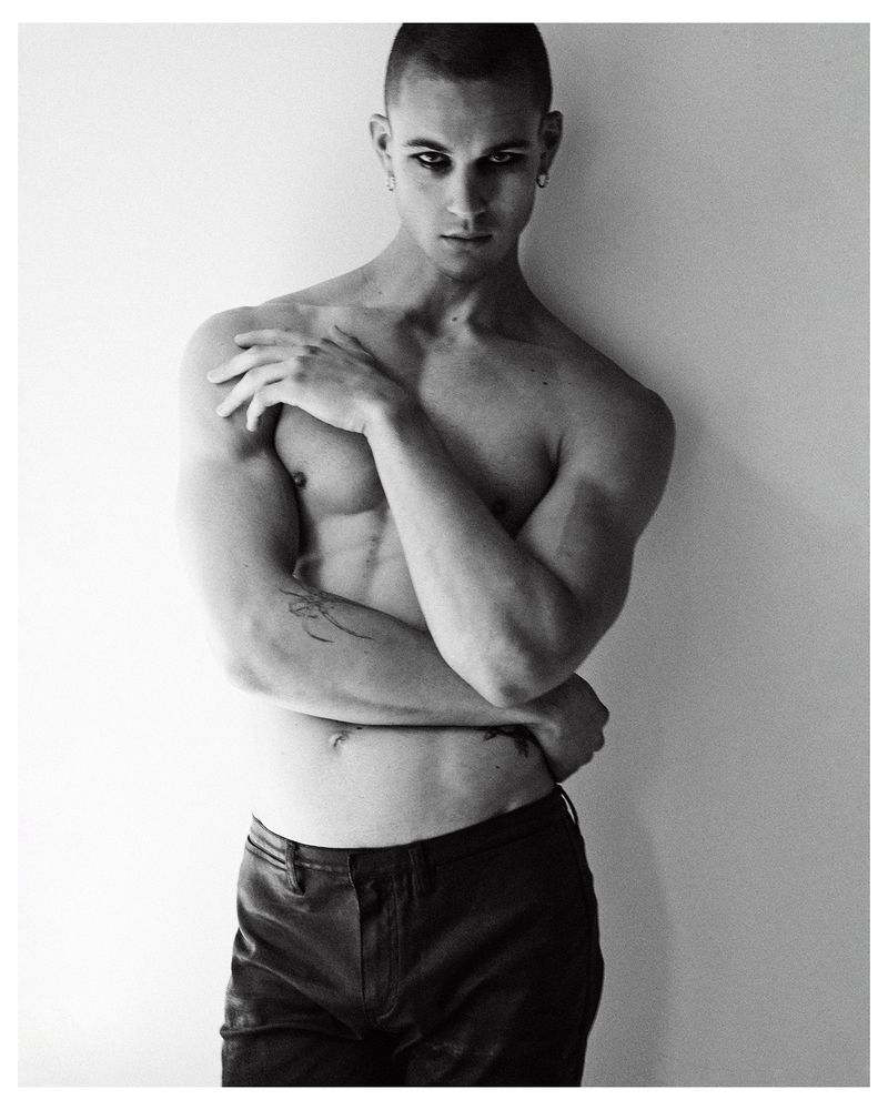 Black and white portrait of a shirtless male model wearing dark pants posing against a plain light wall