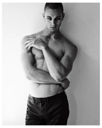 Black and white portrait of a shirtless male model wearing dark pants posing against a plain light wall