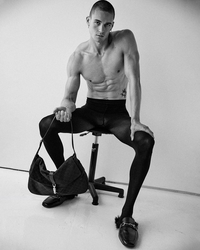 Model Samuel Watson seated on a stool wearing black tights and holding a dark handbag against a plain white background.