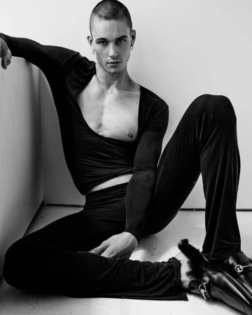 Model Samuel Watson seated against a plain white wall wearing a black top and trousers with fur-trimmed loafers for SCMP Style.