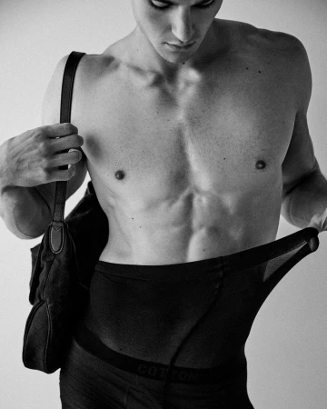 Close-up black and white portrait of male model Samuel Watson holding a shoulder bag while wearing fitted trousers.