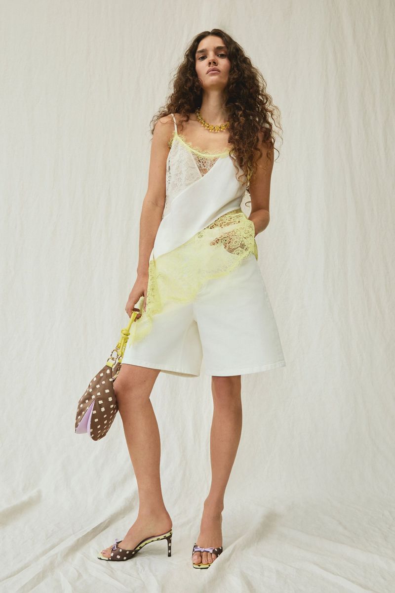 Model in white lace camisole and white shorts holding a patterned brown handbag against a white seamless backdrop.