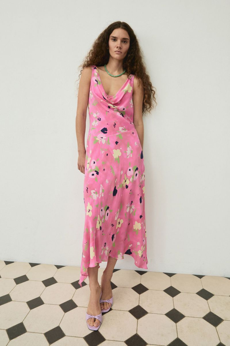 Model with long curly hair wearing a pink floral slip dress against a white wall with a geometric tiled floor