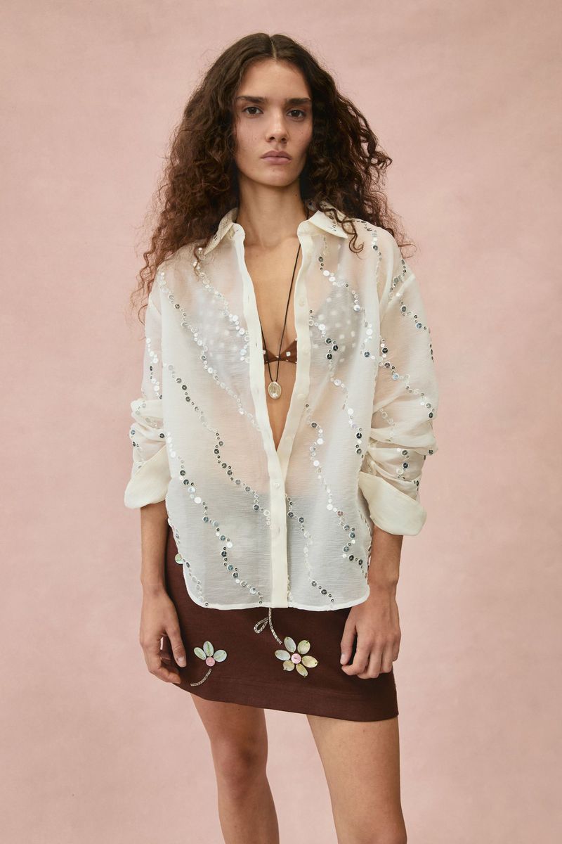 Model in a sheer sequin-embellished shirt and brown floral skirt against a textured pale pink backdrop