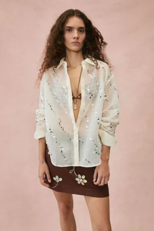 Model in a sheer sequin-embellished shirt and brown floral skirt against a textured pale pink backdrop