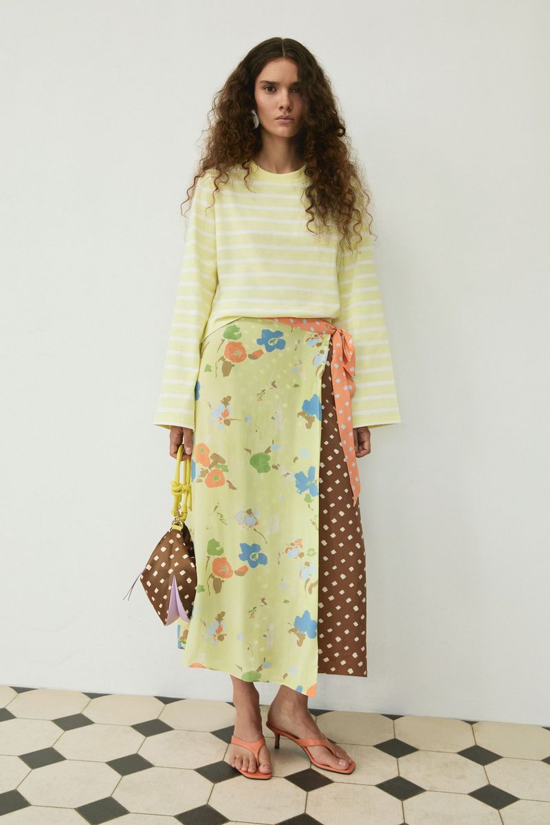 Model wearing a light yellow striped top and patterned floral wrap skirt holding a brown handbag against a white studio wall.