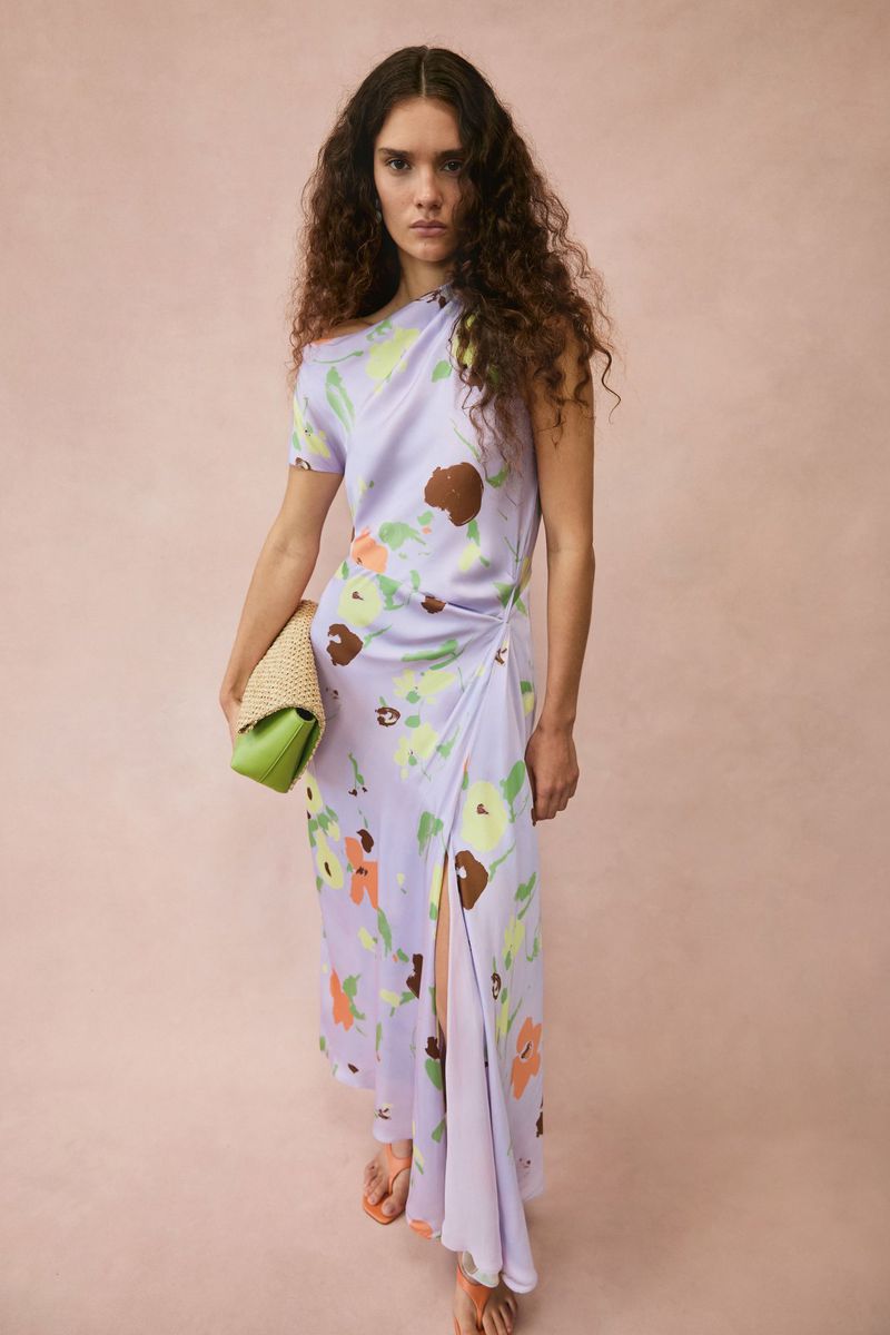 Model with long curly hair wearing a lavender floral print dress and holding a woven clutch bag against a pink studio backdrop.