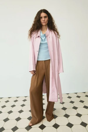 Model with long curly hair wearing a pink coat, light blue top, and brown trousers standing on a patterned floor