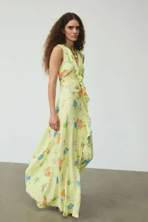 Model with long curly hair wearing a sleeveless yellow floral print maxi dress against a neutral grey studio background