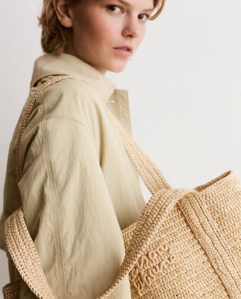 A woman wearing a tan coat poses with a straw tote bag against a light background in a fashion-focused portrait with diffused lighting.