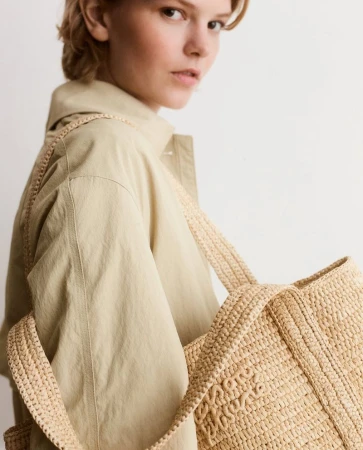 A woman wearing a tan coat poses with a straw tote bag against a light background in a fashion-focused portrait with diffused lighting.