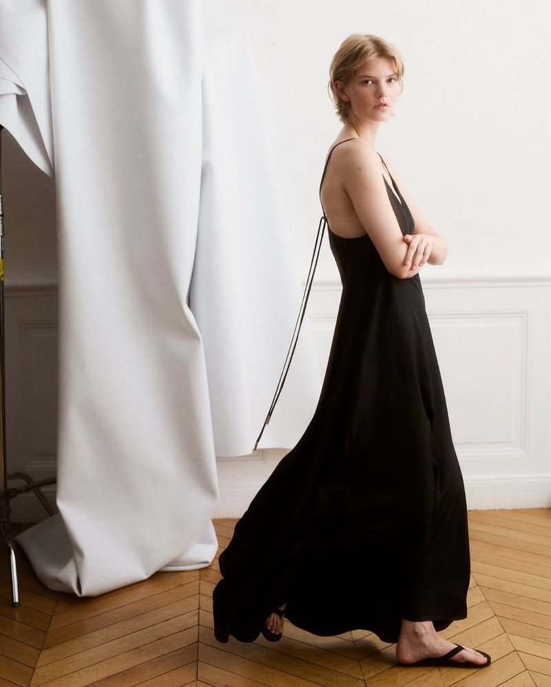 A woman with short blonde hair poses in a black maxi dress in a bright, minimalist studio. She has her arms crossed and looks at the camera.