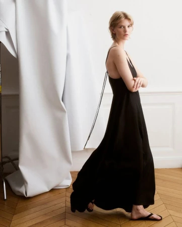 A woman with short blonde hair poses in a black maxi dress in a bright, minimalist studio. She has her arms crossed and looks at the camera.