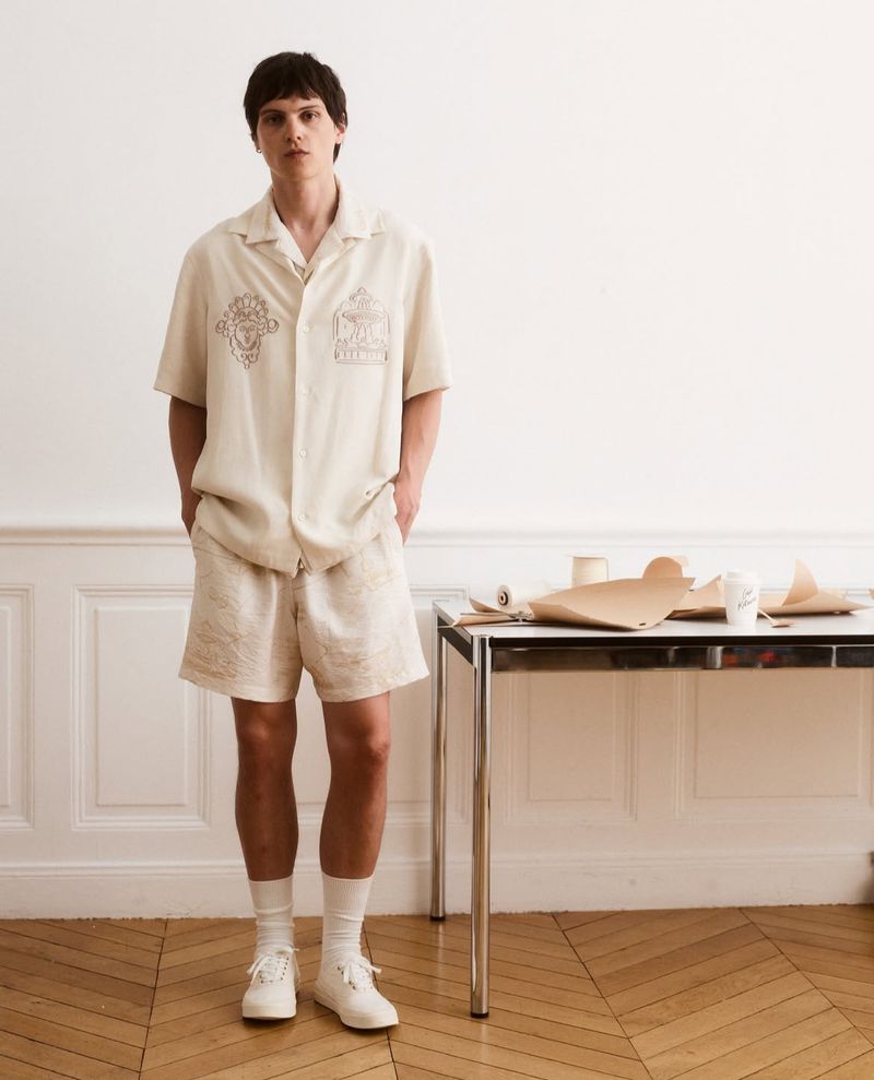 Model Joseph Signoret standing in an off-white embroidered short-sleeve shirt and matching shorts beside a metal table on parquet flooring.
