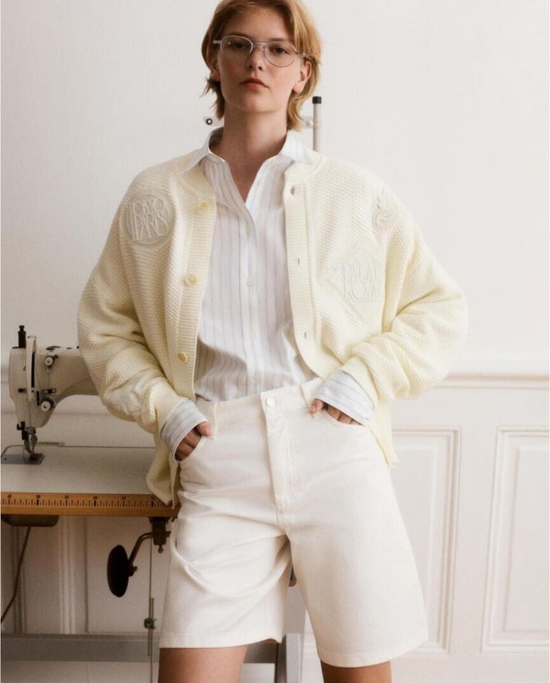 Woman in a light-yellow knit cardigan, white shirt, and white shorts, standing indoors near a sewing machine and a white wall.