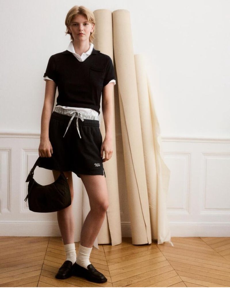 Young woman in a black polo shirt and shorts stands in front of rolled-up beige canvas, posing with a purse and black loafers, in studio.