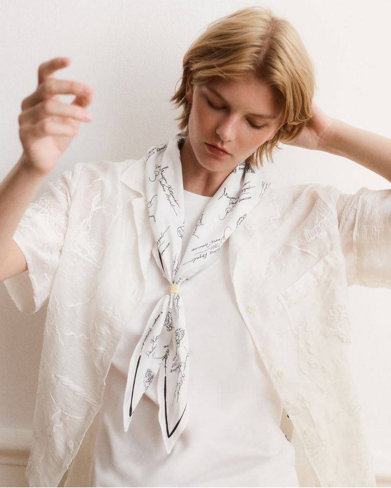 A fair-skinned young adult female in a white button-down shirt poses with a white printed scarf indoors against a neutral background.