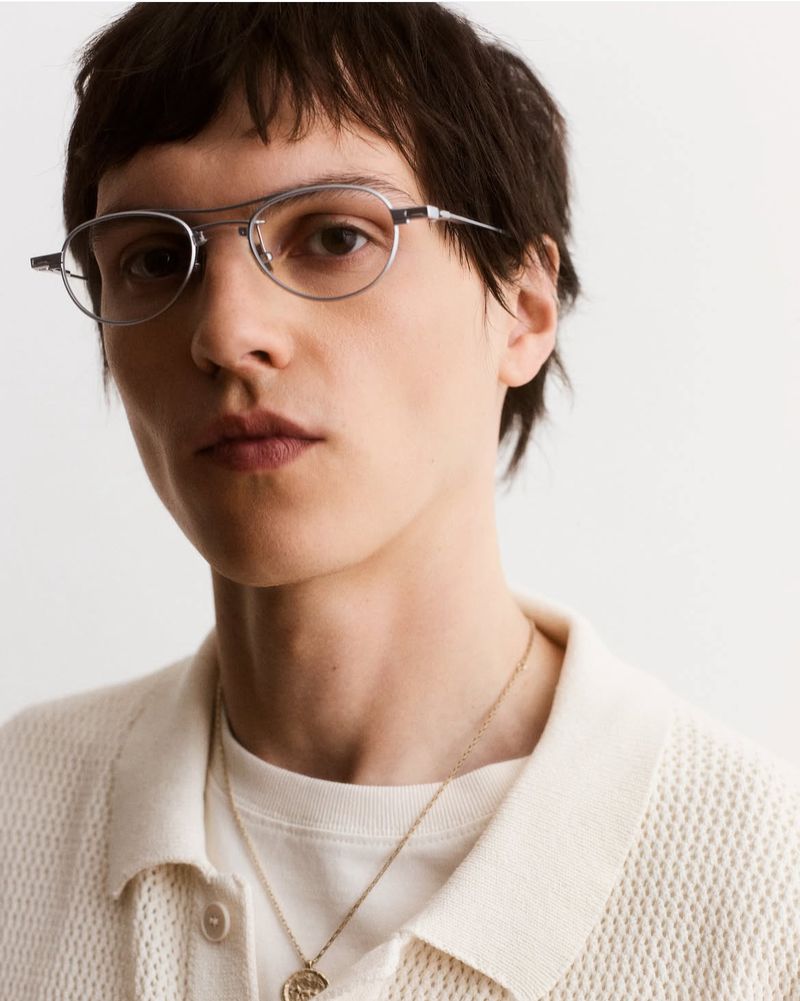 Close-up studio shot of an adult male with dark hair and glasses, wearing a white knit polo shirt with a gold pendant necklace.