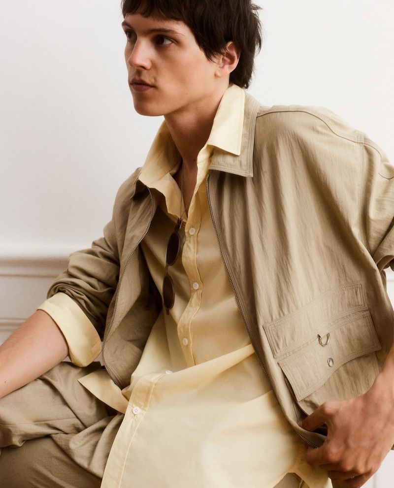 Tan-skinned male model with short dark hair poses indoors wearing a yellow button-down shirt with a tan jacket and matching pants.