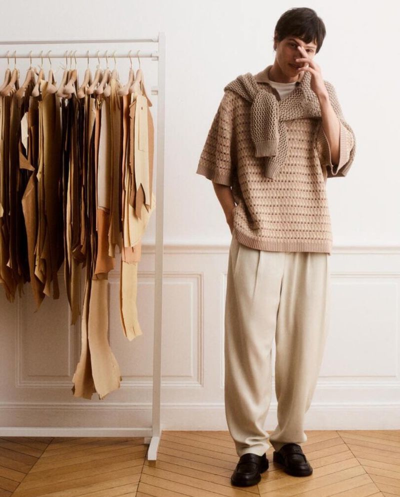 Man in beige sweater and loose pants stands near a rack of fabric swatches in a minimalist indoor setting. Fashion editorial shot.