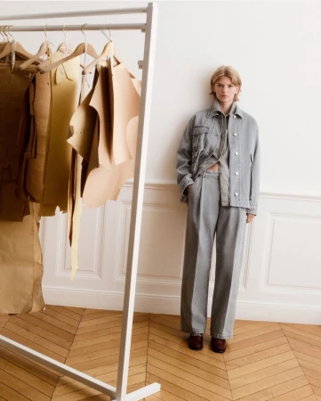 Indoor shot of a fashion model posing in an outfit, alongside a clothing rack with garment patterns in a minimalistic setting.