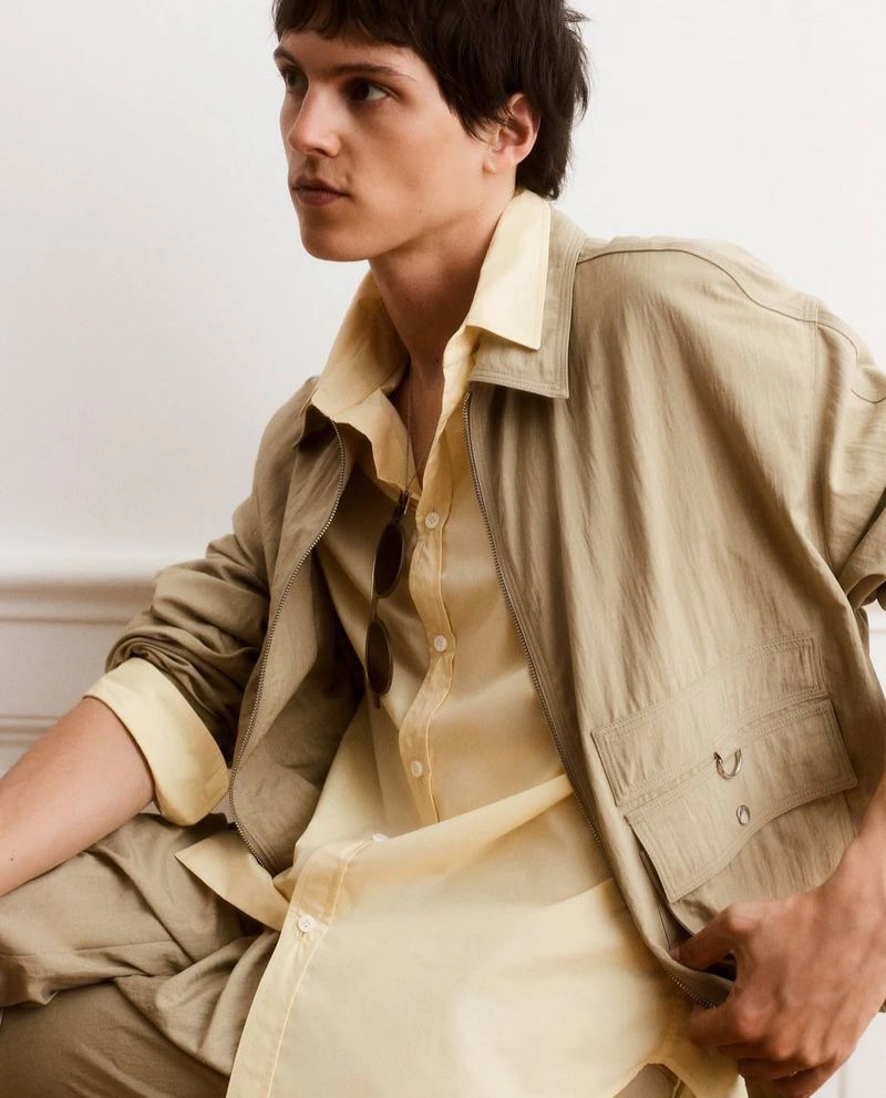 Young male model wearing a beige utility jacket over a yellow shirt against a plain wall.