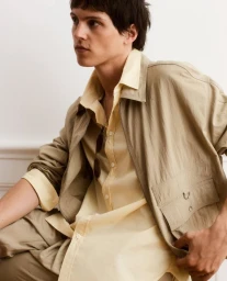 Young male model wearing a beige utility jacket over a yellow shirt against a plain wall.