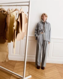 Model wearing a grey denim jacket and matching trousers standing beside a clothing rack in a light room.