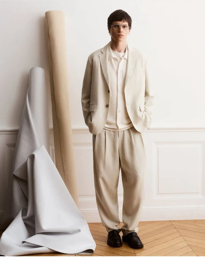 Model standing in neutral tailored suit jacket and trousers against a wall with draped fabric rolls for Maison Kitsuné