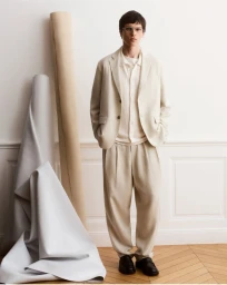 Model standing in neutral tailored suit jacket and trousers against a wall with draped fabric rolls for Maison Kitsuné